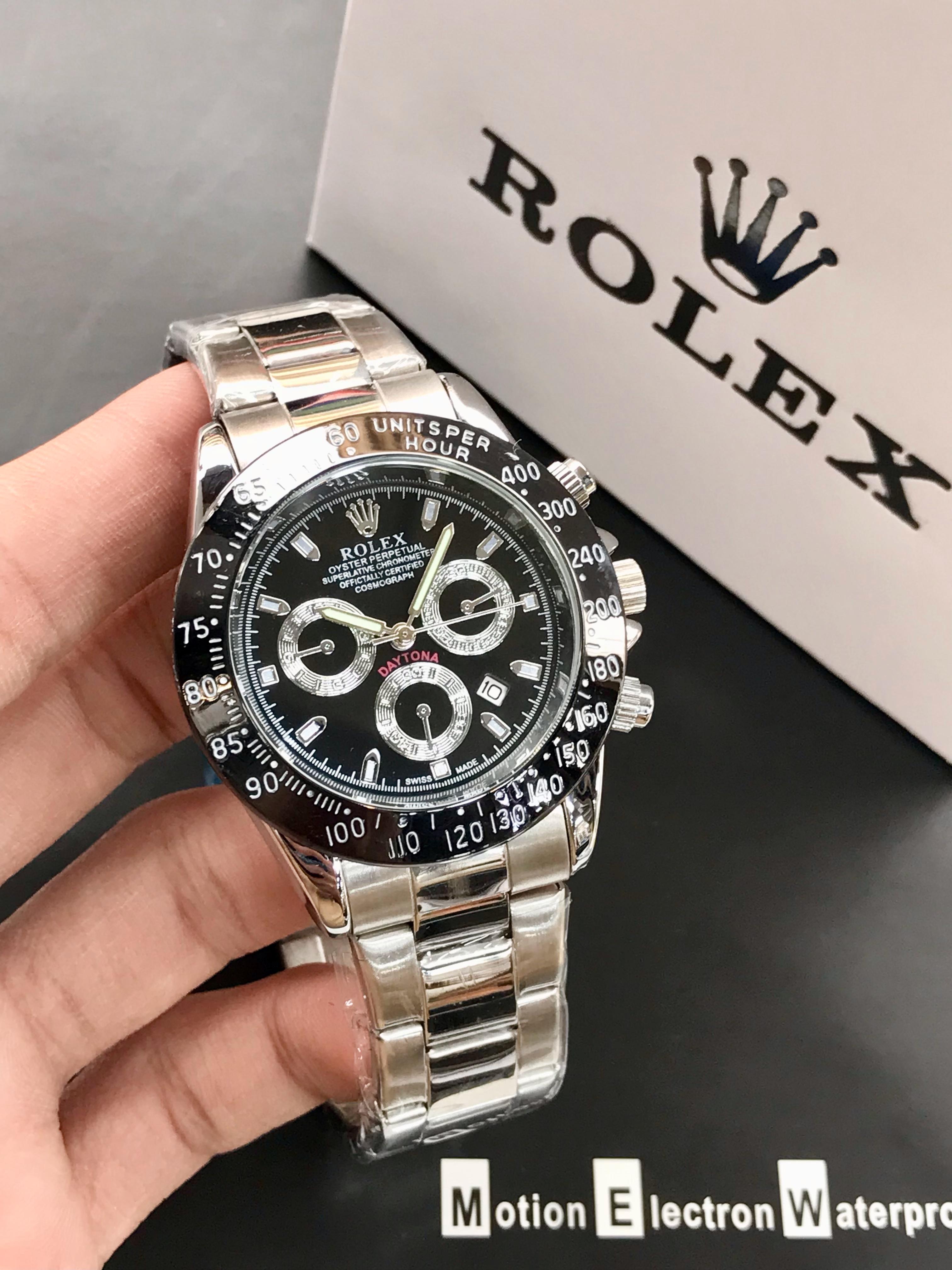 Silver and black Rolex watch held by a hand with a Rolex box in the background.