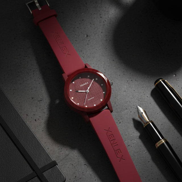 Red watch with 'Xenlex' branding on a dark surface with a pen and ink bottle.
